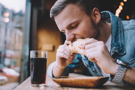 Young attractive man sitting in a cafe near window during the break. Nice guy is in the process of eating burger and drinking cold tea or juiceの写真素材