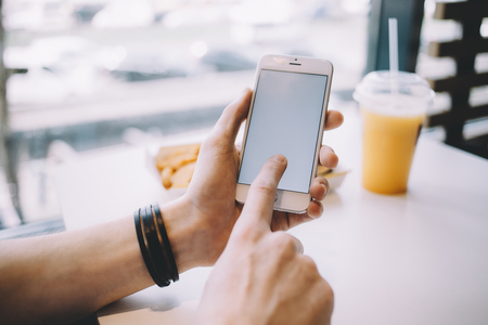 Young mans hand holding mobile phone. Stylish guy sitting in cafe and using his gadget scrolling some stuff. Place for your advert or logo. Back view. Close up. Horizontal position of the phoneの写真素材