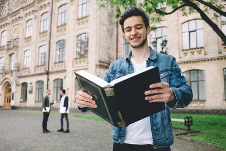 Smiling student in jeans jacket reading a huge book in front f the camera and holding it with both hands standing near the college. Close upの写真素材