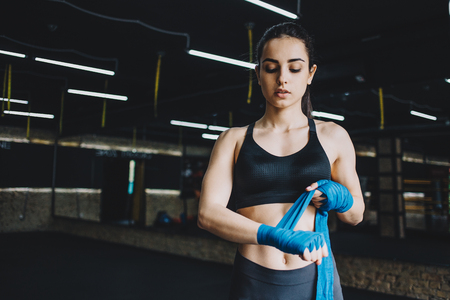Beautiful sporty woman wrapping her fists with blue bandage tape. Fit girl is standing in gym and preparing to the fight. Close upの写真素材