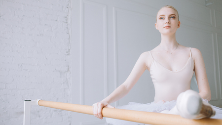 Beautiful ballerina standing near barre and putting her leg on it in a classical ballet position. She is dreaming about being famous and dancing at big worlds stages. Cut view. Close upの写真素材