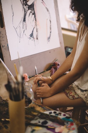 Talented artist girl with black curly hair painting picture on canvas in studio. Cute woman using different brushes and paints for her workの写真素材