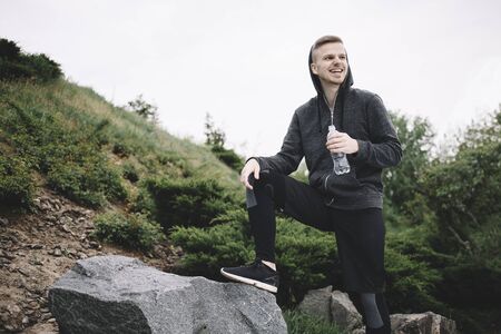 Young jogger has put his right leg on the stone and holding a bottle of water in the left hand. He decided to have a short break. This guy looks happy and smilingの写真素材