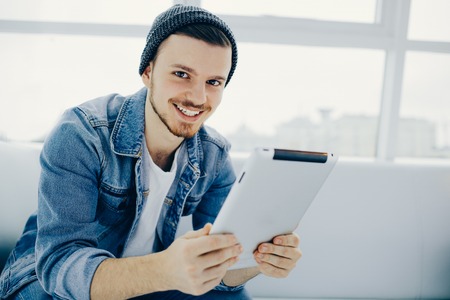 young, successful business man sitting in cafe with tablet on sofa. Serious student is looking at camera holding his tablet and smilingの写真素材
