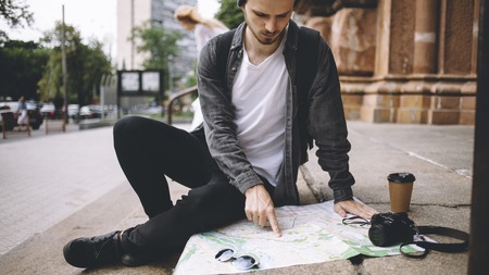 Close view of a young hipster that likes to travel. He decided to have some rest and sit down on the cathedrals steps on the street. His coffee is getting colder while the guy is stading the map of the city. Close upの写真素材