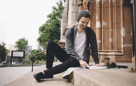 Happy and smiling tourist is sitting on the churchs steps on the street. He took a cup of coffee and decided to have some rest. Now he is studing the map of the city. His camera is placed near the cup of coffee.の写真素材