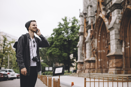 The photo of a traveller standing on the sidewalk near the old church in a big town. He is standing toward the building and talking on the phone. He is smiling to himself and looking straight forward.の写真素材