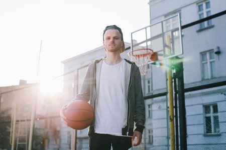 The portrait of a young man that stands near the basket in a bright sunny day. He is holding the ball for the basketball game with his right hand. He is looking straight to the camera.の写真素材