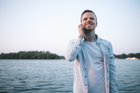A beautiful evening picture of a young nice man stayung near the big river in the evening. He is talking on the phone and looking straight ahead. This man is happy. Close upの写真素材