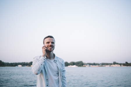 A cut view of a young guy standind near the big river and talking on the phone. He is enjoying the conversation so thats why he is smiling. This man looks happy.の写真素材