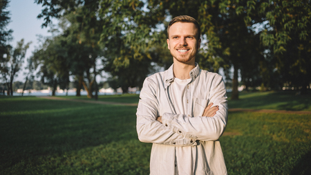 A portrait of a happy successful student standing in a park and posing to camera. His lessons are over so now he has some free time to spend outside and enjoy the warm weather and evening sunlight. The young man is smiling to the camera.の写真素材