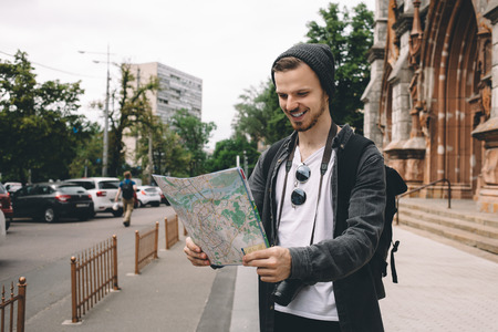 Curious and ambitious young tourist is standin on the street and holding a city map in his hands. He...