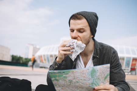 Good-looking and attracive tourist is sitting on the big white steps. He is eating a burger with his right hand and looking to the map which he is holding with his left hand. He is searching an interesting place to visit. Close up. Cut viewの写真素材