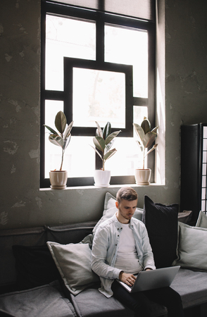 Vertical picture of a guy sitting with a laptop on the couch near the big bright window. Young man is chatting with another gamers and trying to find good players to play with.の写真素材