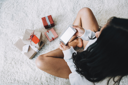 Another view of a girl sitting on the white furry carpet and holding a phone in his hands. She wants to take a picture of Christmas presents that are lying on the carpet. All of them are small but pretty. Close up. Cut viewの写真素材