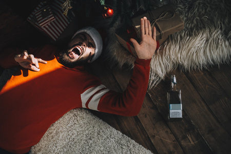 One adult is lying on the floor near pine tree in room in the sunny morning. He is trying to cover his face from the sunlight. Plus this guy has a morning hangover after drinking too much alcohol last evening during the Christmas celebration. Cut viewの写真素材