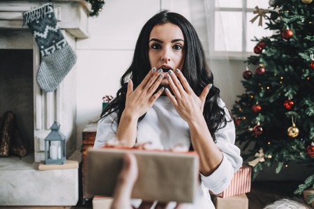 Emotional picture of a beautiful girl getting a present. She is sitting on white furry carpet and looking straight to the camera. It is so unexpected for her to get a present. She is really amazed. Cut viewの写真素材