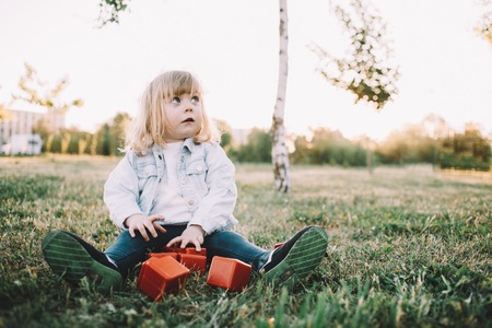 Small and cute baby-girl is sitting on the grass and looking to the right. She is touching some orange cubes that she has besides her legs. Her sight is full of curiosity.の写真素材