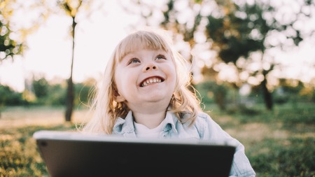 Nice picture of a young girl sitting on grass and looking up with a kid smile. She is holding a big tablet in her hands. The child wanted to watch a cartoon when suddenly something interesting appeared in the sky. Close up. Cut viewの写真素材