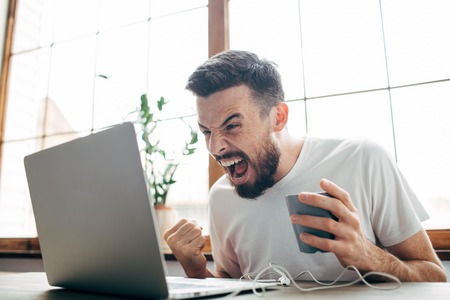 Angry man is sitting in the room near big window and looking to the laptops screen. He is angry. Guy is screaming to the screen. Also he is holding a cup of tea in the left handの写真素材