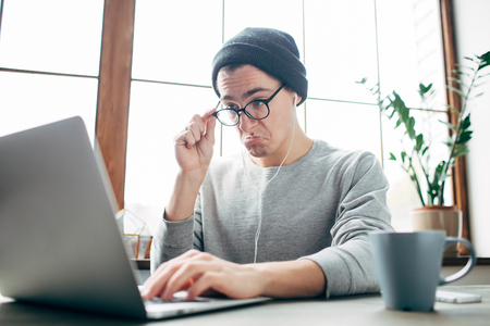 Guy is working at the table. He is looking to the laptop with wonder. He has almost put off his glassesの写真素材