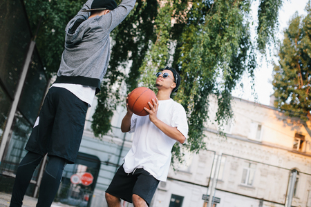 Two guys are training. The boy in white shirt is getting ready to throw the ball while the afroamerican guy is defencing the basket and trying to prevent ball hit the basketの写真素材