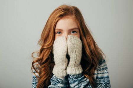 The girl in a sweater with red horns smiles, warming her face with gloves on her hands. Warm knitted gloves turned out to be very warm and warmly warm her hands. Isolated white backgroundの写真素材
