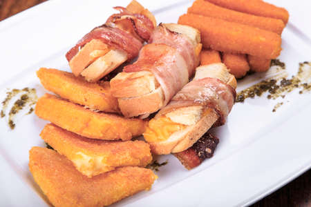 a plate of fried snacks on the table. in a plate are rolls of bacon, fried cheese, breaded, fried chicken snacks. close-upの写真素材