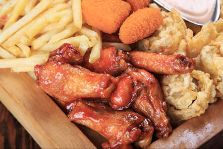 wooden board on a table with assorted fried appetizers: BBQ wings, fried squid rings, fries, grilled cheese, sauceの写真素材