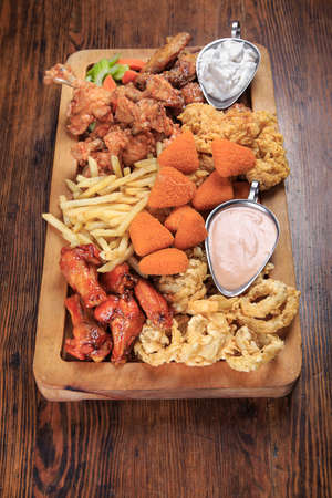 wooden board on a table with assorted fried appetizers: BBQ wings, fried squid rings, fries, grilled cheese, sauceの写真素材
