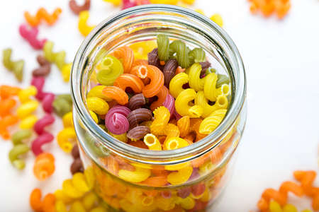colored pasta in a jar and some scattered on the white wooden tableの写真素材