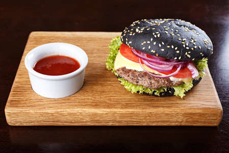 Black burger on the board with fried potatoes; cutlet; vegetables and cheese; with sauce on a wooden boardの写真素材