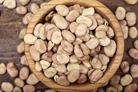 Dried broad beans in a wooden bowl on a wooden background. Next to a plate, beans are scattered on the tableの写真素材