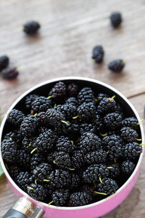 black mulberry berries in a pink pot on a wooden table. in the foreground are mulberry berries and...