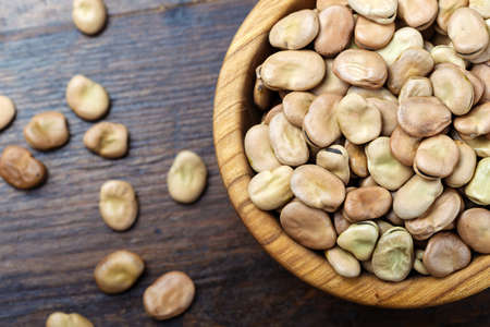Dried broad beans in a wooden bowl on a wooden background. Next to a plate, beans are scattered on the tableの写真素材