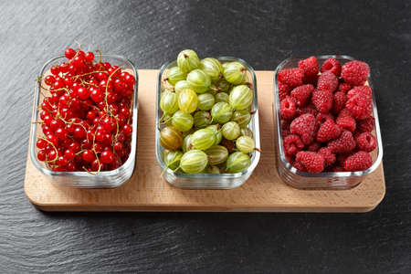 Raspberry, red currant and gooseberries, mix of summer berries in the same glass plates on a wooden stand on a black slate background, top viewの写真素材