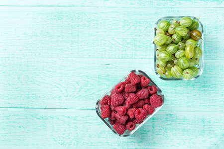 Raspberries and gooseberries, mix of summer berries in identical glass plates on a wooden stand on a wooden turquoise background, top viewの写真素材