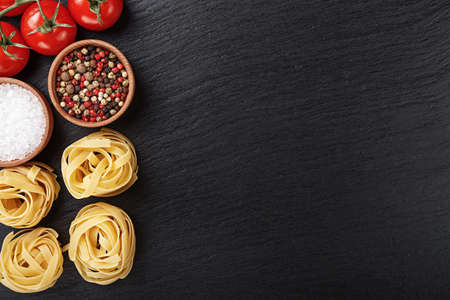 Pasta tagliatelle on a black background, next to lie raw tomatoes, salt and pepper. place for textの写真素材