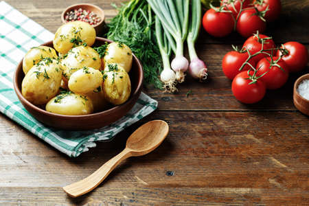 A young potato with dill in a clay plate on a wooden table, on the table there are onion, spices, fresh tomatoesの写真素材