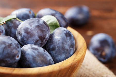 ripe plums in a wooden plate, with leaves, on a wooden background