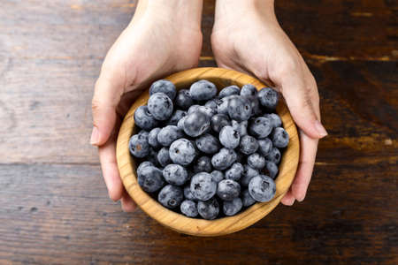 ripe blueberries in a wooden plate, in hands, on a brown wooden background. clearly visible texture of berriesの写真素材