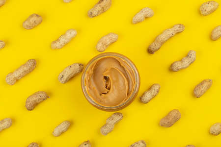 Bowl of peanut butter and scattered peanuts on yellow background, top view.