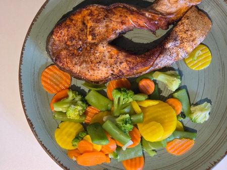 Tasty grilled salmon steak served with a colorful mix of steamed vegetables including carrots, broccoli, green beans, yellow carrots, and cauliflower on a ceramic plate. Healthy balanced meal, rich in protein and nutrients. Ideal for diet, fitness, and clean eating concepts.の写真素材
