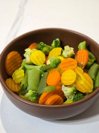 Healthy bowl of mixed steamed vegetables including broccoli, carrots, yellow carrots, cauliflower, and green beans. Colorful vegetarian meal in a brown bowl on a white table. Clean eating, vegan food concept.の写真素材