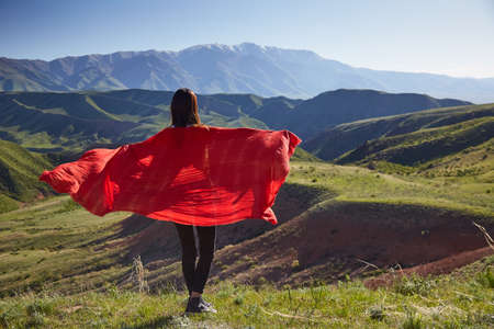 A girl with a red wrap on her shoulders against the background of the spring mountain landscape. View from the back. Mountain spring landscapeの写真素材