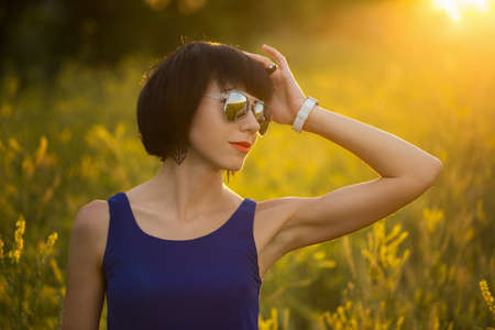 Portrait of a girl at sunset on the background of a blooming field of yellow flowers. Girl in sunglasses and a purple dress. Summer moodの写真素材