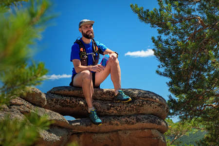 A male runner is sitting on the edge of a cliff, he is resting after a successful workout. Outdoor trail running. Man in blue t-shirt and black shortsの写真素材