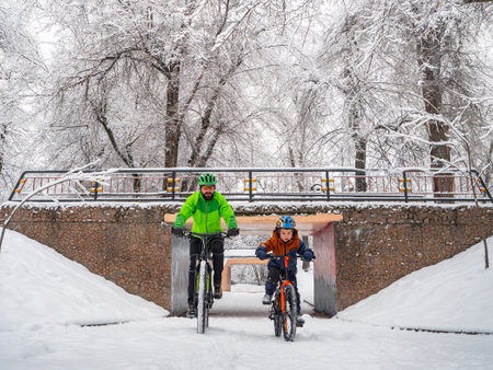 A boy and his father ride bicycles in a winter park. Wonderful winter day outdoors after snowfall. The bearded man is the boy's father, follows him on a bicycle.の写真素材