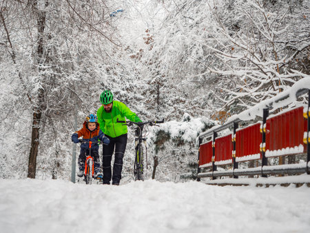 Father teaches his little son to ride a bike in winter. An active pastime in the fresh air after a snowfall in a winter park.の写真素材