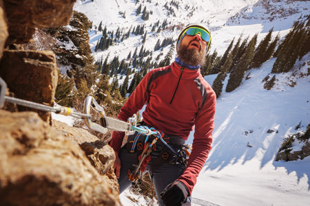 Bearded climber, secured with a belay device, looks at the top of the cliff. Winter alpine mountaineering. A man climbs a rock. Mountain climber portraitの写真素材